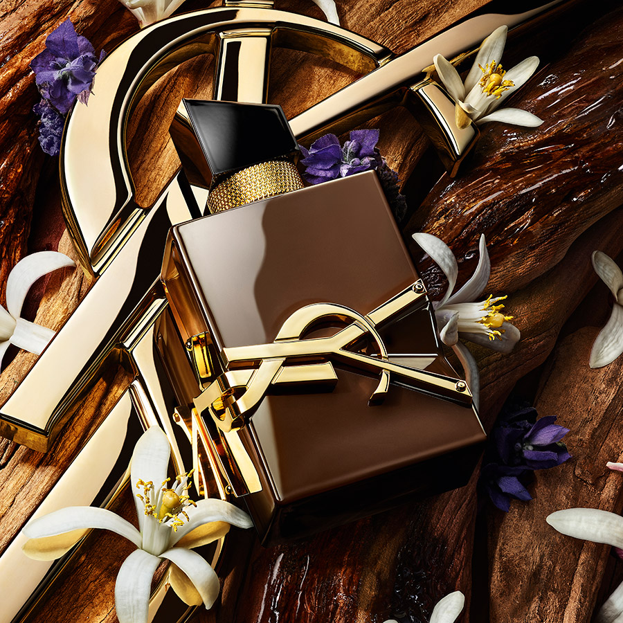 Un flacone marrone del profumo YSL Libre appoggiato sul legno accanto a un logo dorato, fiori d'arancio bianchi e lavanda viola.