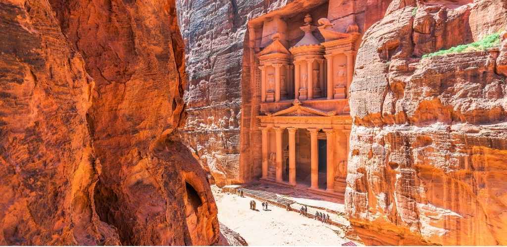 The Treasury (Al-Khazneh) rock-carved facade in Petra, Jordan, framed by sunlit red sandstone cliffs with tourists