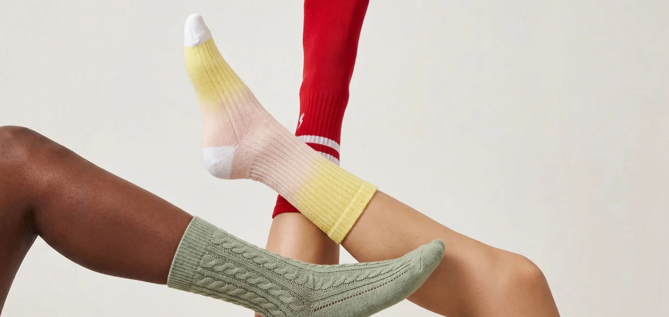 Tre gambe sollevate con calzini colorati: sfumatura verde, rossa e gialla, su uno sfondo chiaro e uniforme.