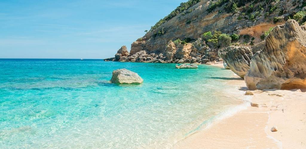Turquoise Mediterranean bay with crystal-clear water, sandy beach and rugged limestone cliffs under a clear blue sky