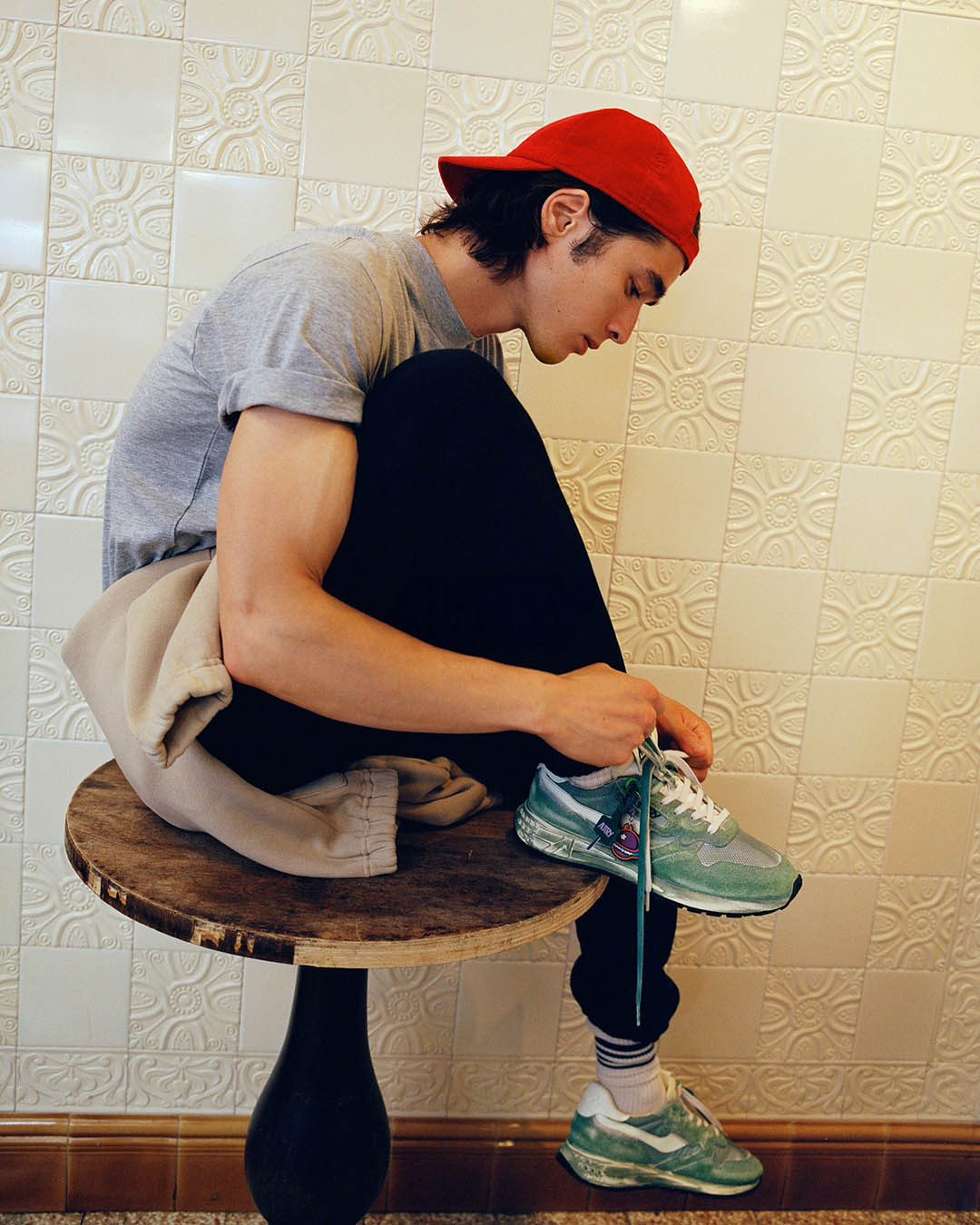 Teenager in red cap sitting on a wooden stool tying green trainers' laces against a decorative tiled wall