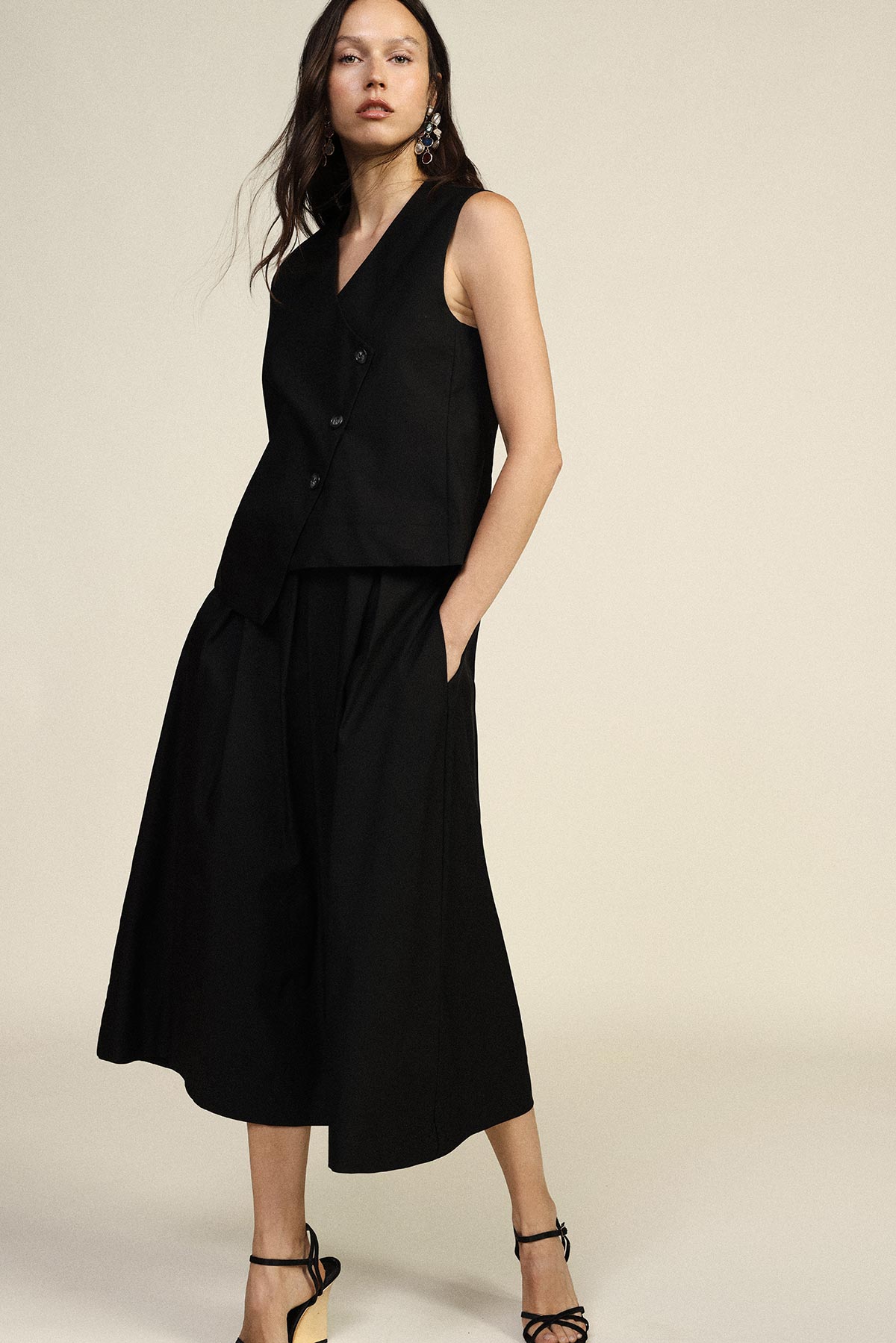 Model wearing sleeveless black tailored vest and wide-leg culotte trousers with strappy black heels on neutral background