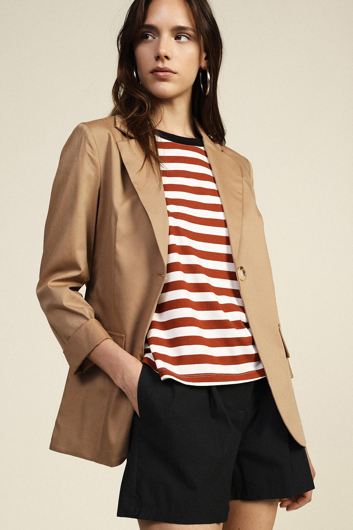 Woman wearing a tan blazer over a red and white striped top and black shorts, hands in pockets, looking to the side