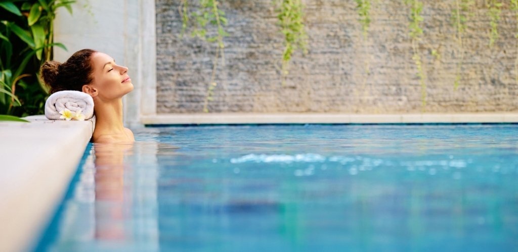 Person relaxing in a spa pool, leaning on the edge with a rolled towel and frangipani flower, tranquil water and textured waterfall wall