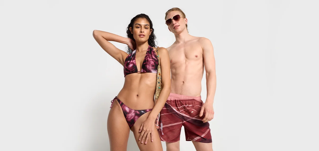 Donna in bikini a tie-dye viola con borsa intrecciata e uomo in pantaloni da bagno rossi a fantasia e occhiali da sole che posano contro uno sfondo bianco semplice.