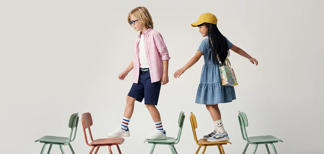 Due bambini si bilanciano e camminano su una fila di piccole sedie colorate distanziate su uno sfondo semplice.