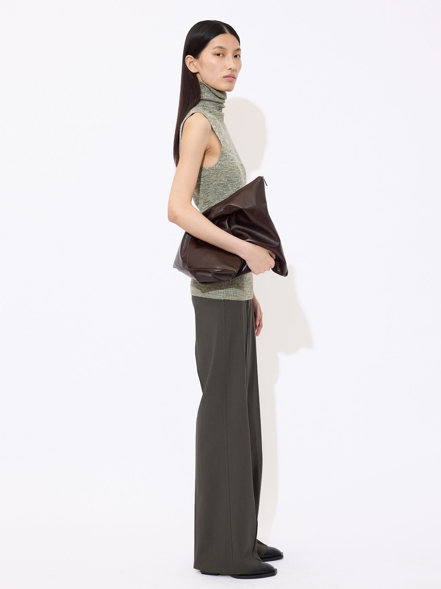 Woman wearing sleeveless green turtleneck and olive wide-leg trousers holding large brown clutch on white background