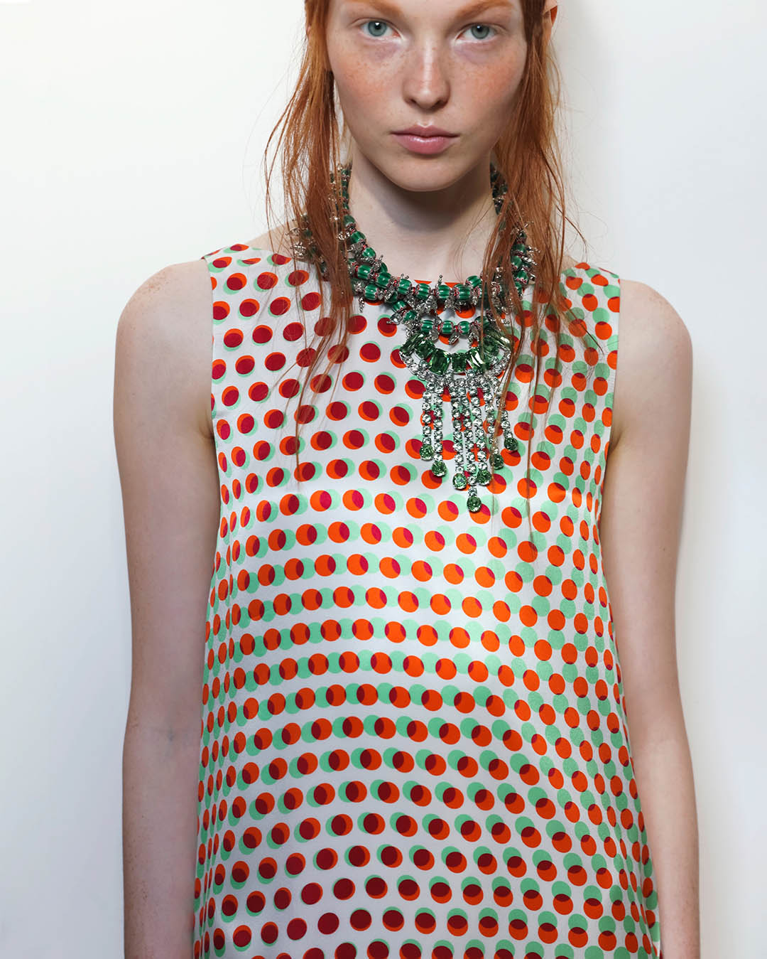 Red-haired model in sleeveless white dress with red and green polka dots and ornate green statement necklace