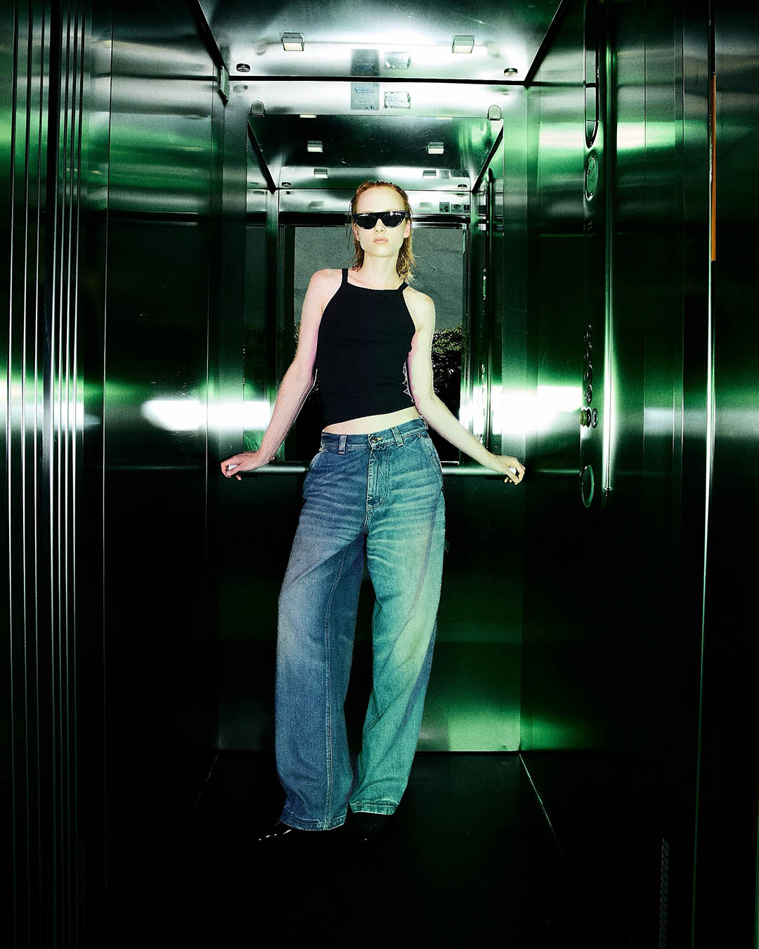 Person in sunglasses posing in a green-lit mirrored elevator wearing black crop top and wide-leg jeans