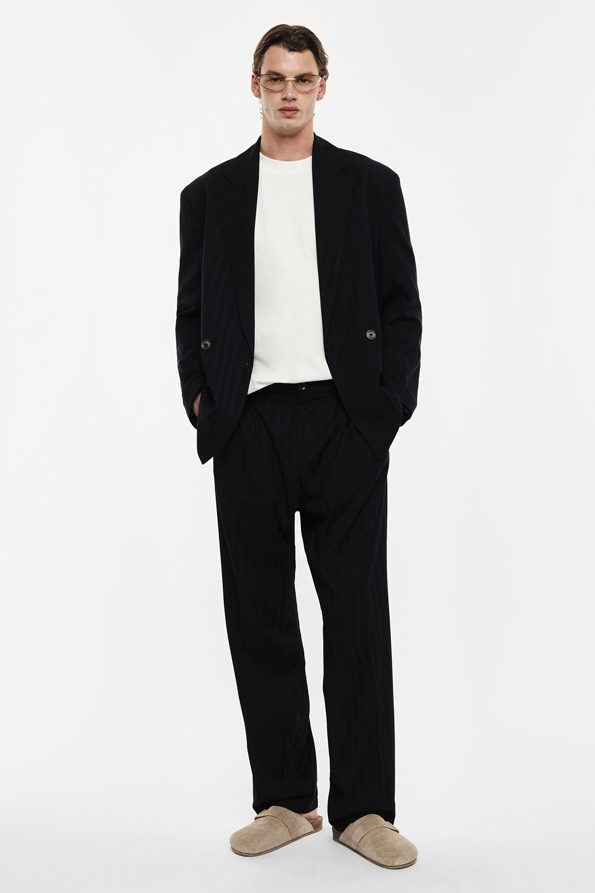 Male model in oversized black suit and white T‑shirt, wearing glasses and beige slippers on plain white background