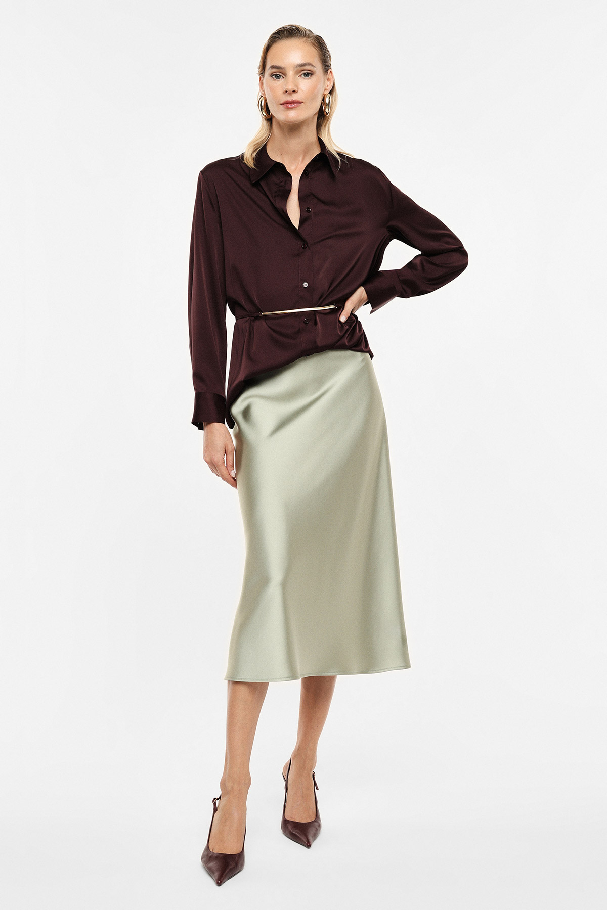 Female model wearing burgundy button-up blouse with slim belt, pale sage satin midi skirt and brown pointed heels, studio
