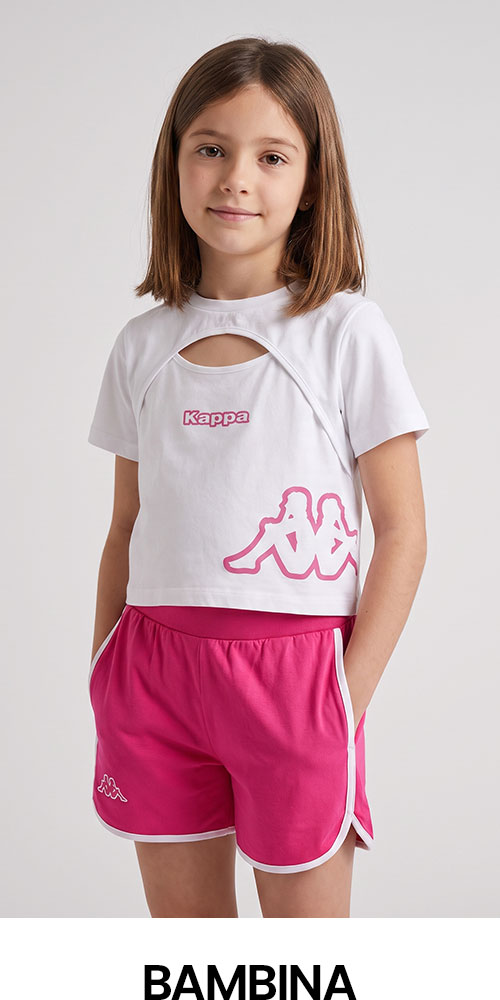 Young girl modelling white Kappa cropped top with pink logo and pink shorts, hands in pockets against plain background