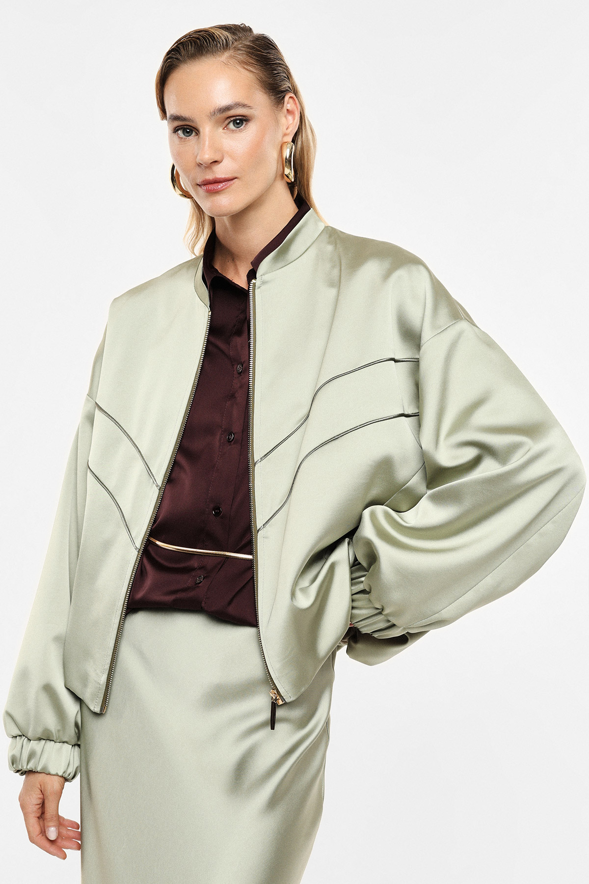 Fashion model wearing sage-green satin bomber jacket and matching skirt over burgundy silk blouse, gold hoop earrings