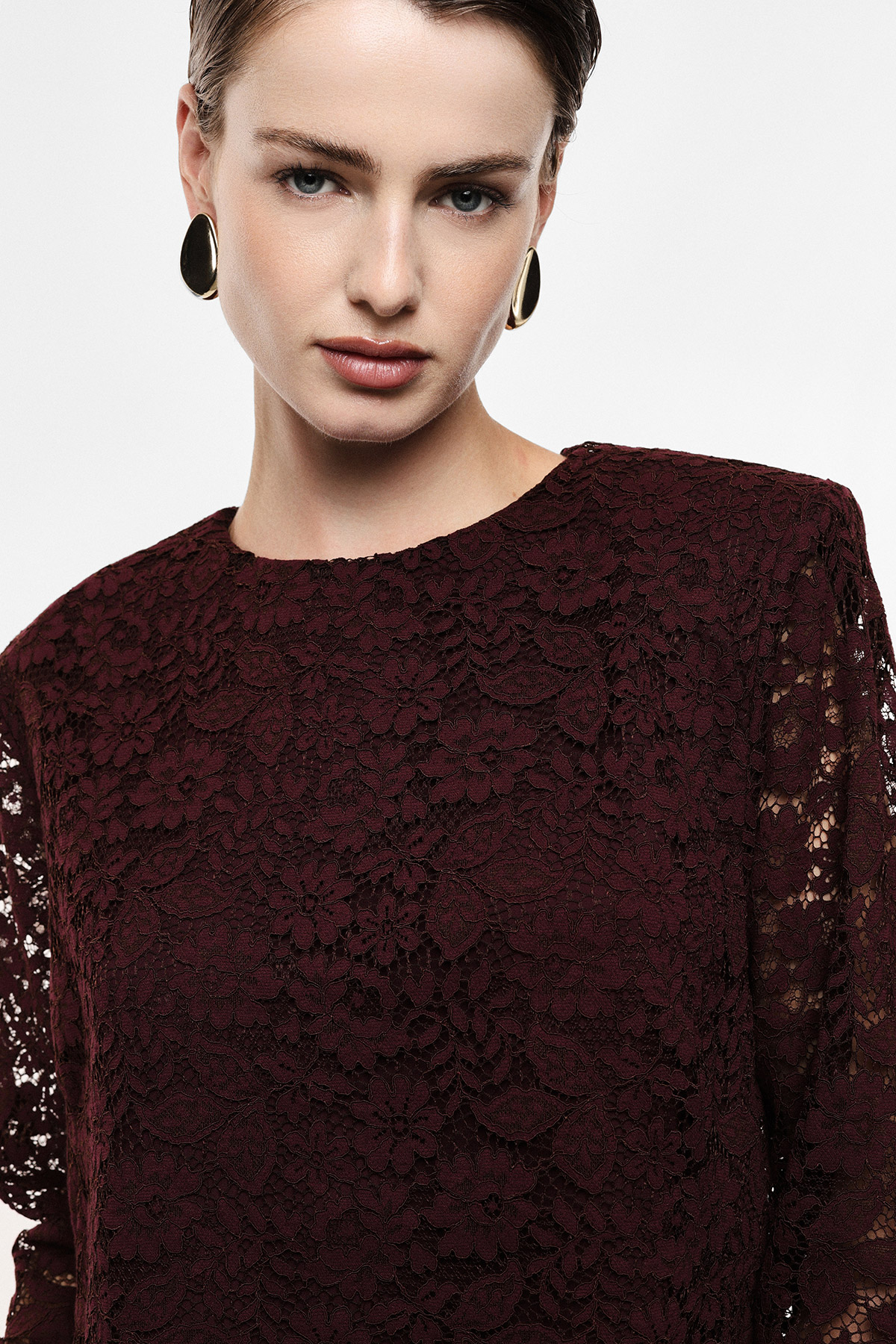 Close-up of woman wearing burgundy lace top and gold oval earrings, neutral makeup, cropped on white background