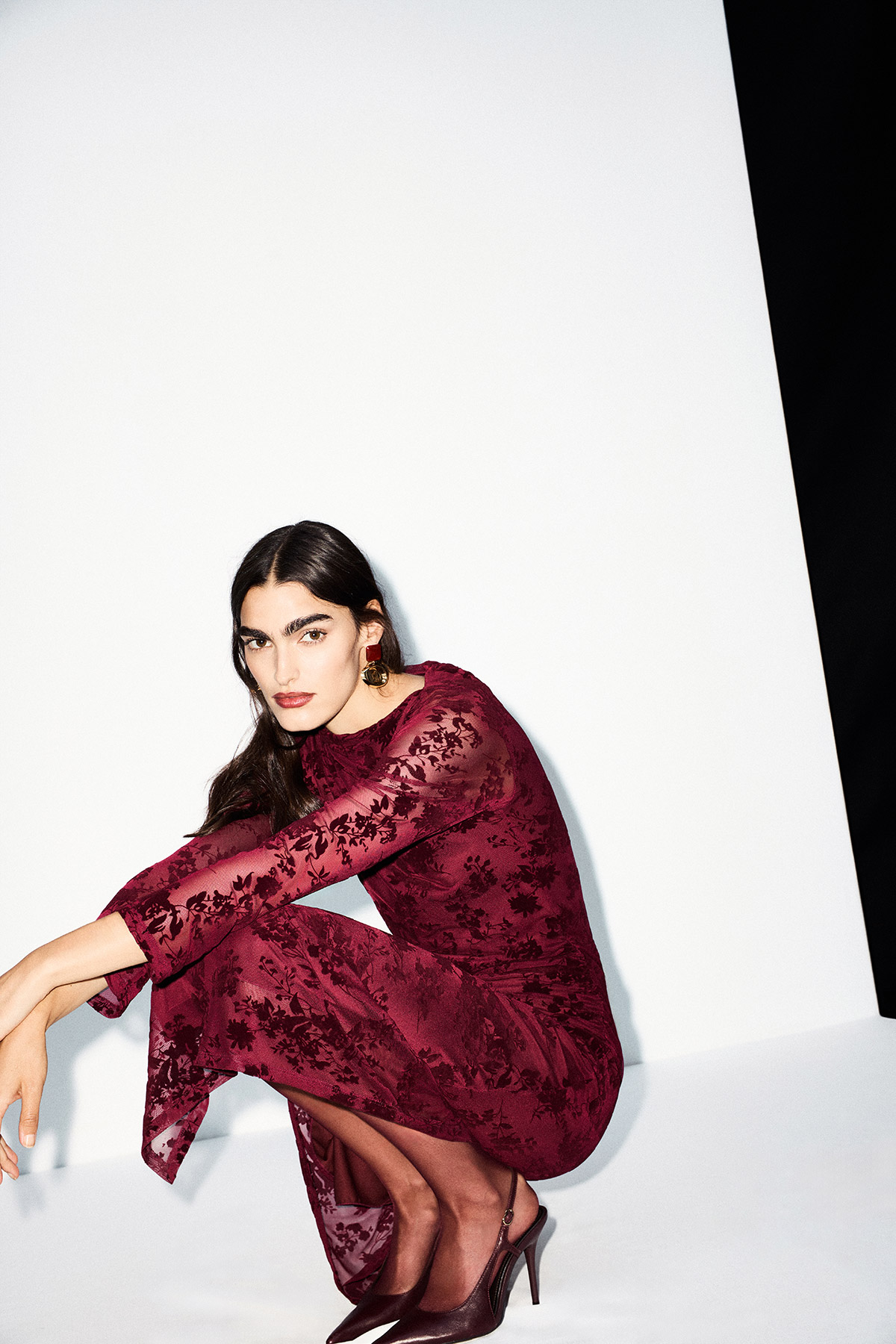 Woman in burgundy lace midi dress crouching against white backdrop, wearing heels and statement earrings — fashion editorial pose