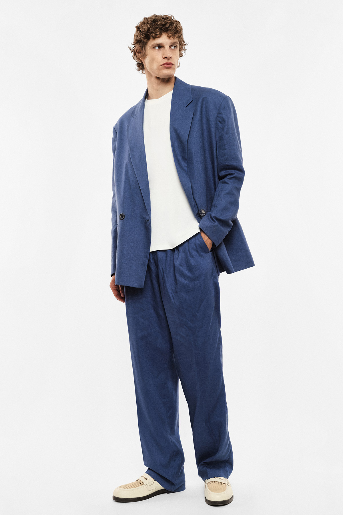 Young man in relaxed blue suit and white T-shirt with cream trainers, hands in pockets, studio fashion portrait