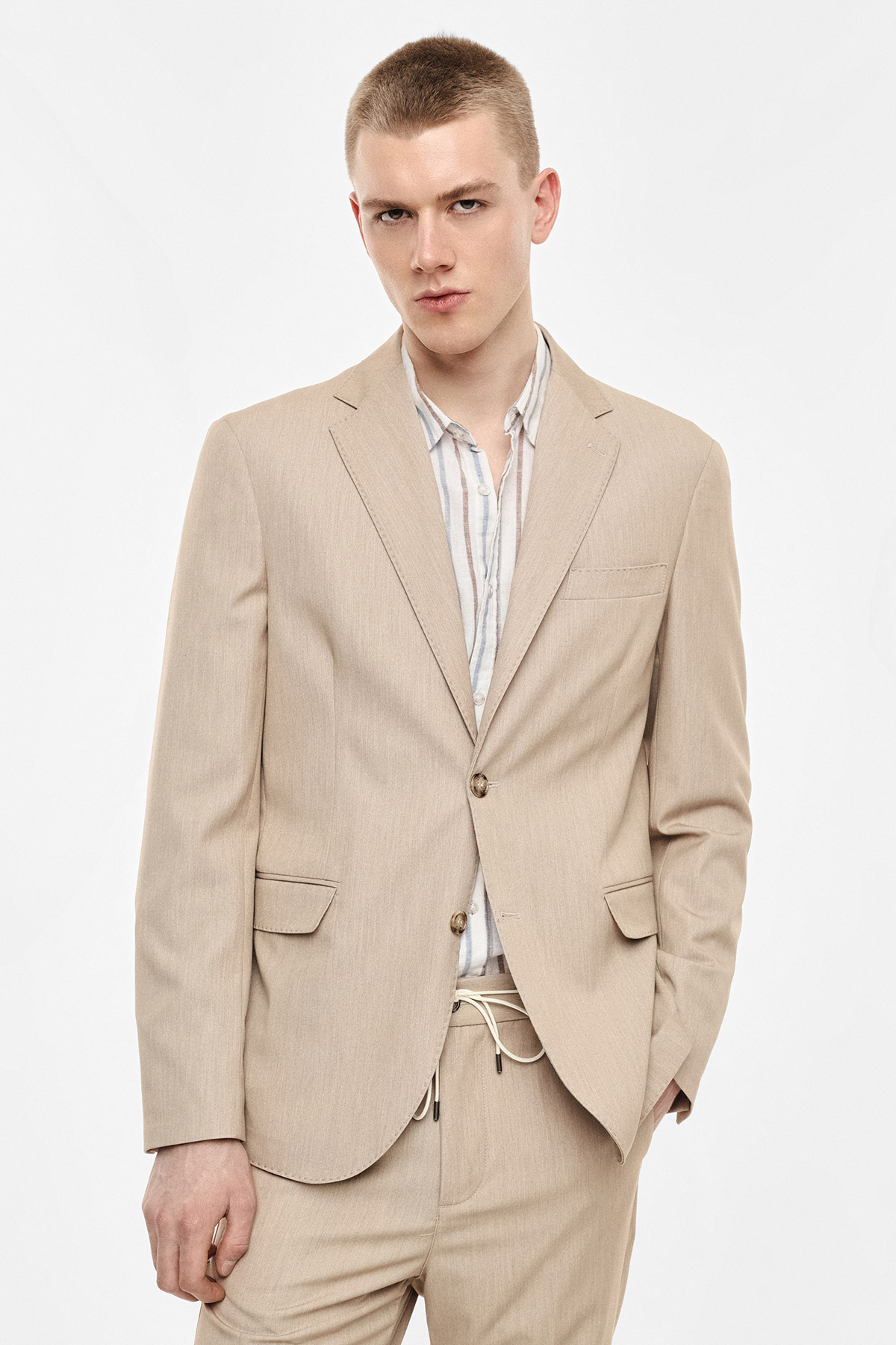 Male model in beige two-piece suit and striped shirt, relaxed pose with hand in pocket against plain white background