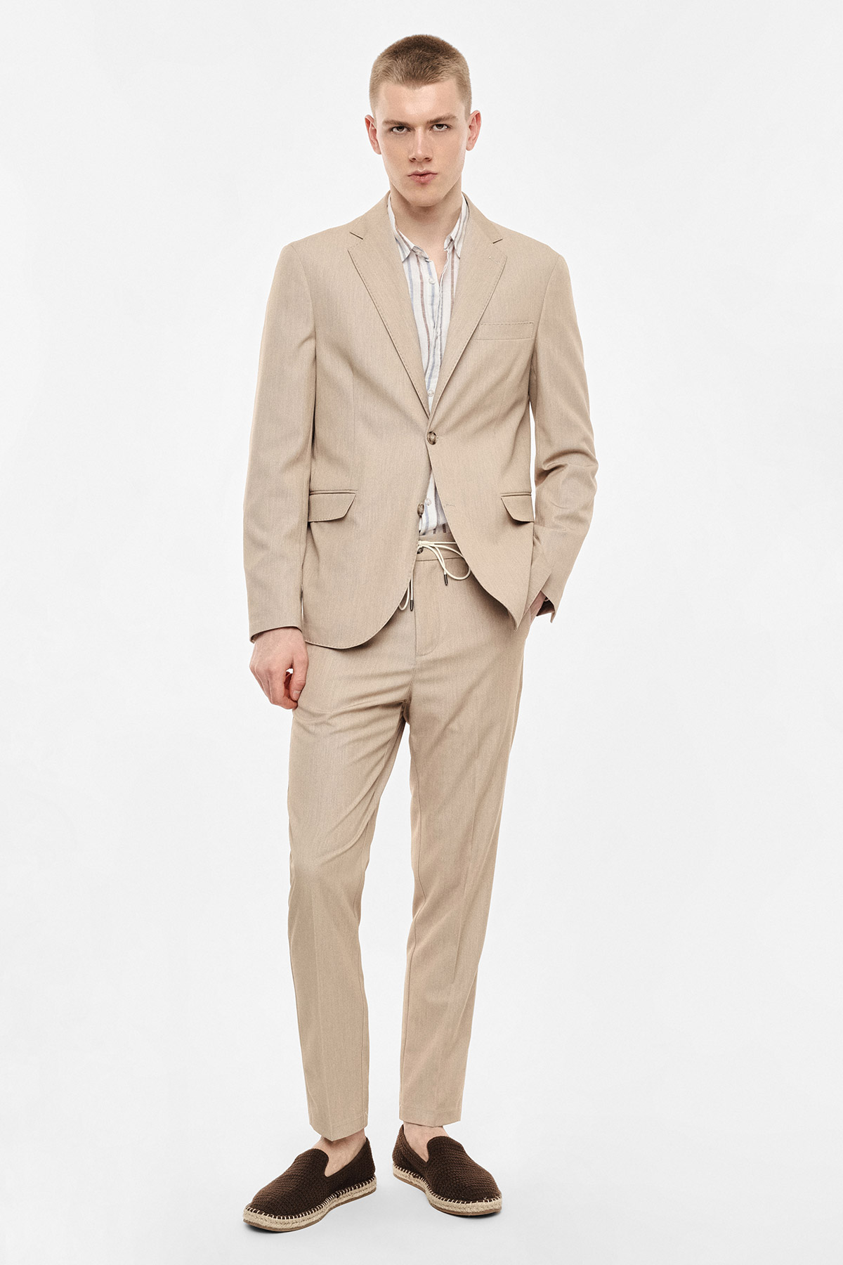 Young man standing in a beige suit with a striped shirt and brown espadrilles, hands in pockets on a plain white background