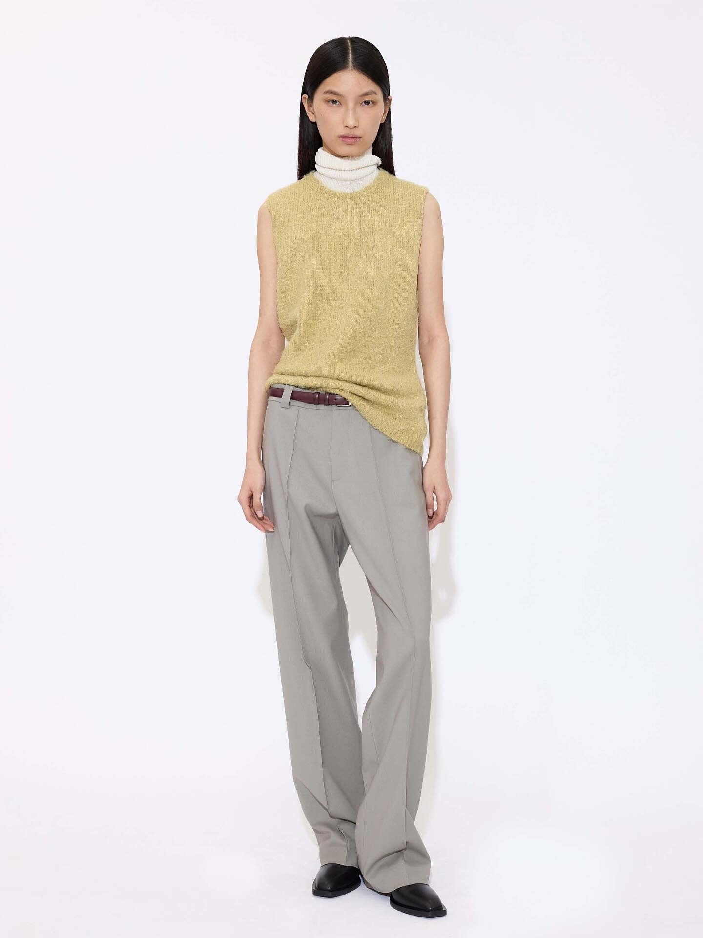 Woman in sleeveless yellow knit over white turtleneck, grey wide-leg trousers, brown belt and black shoes