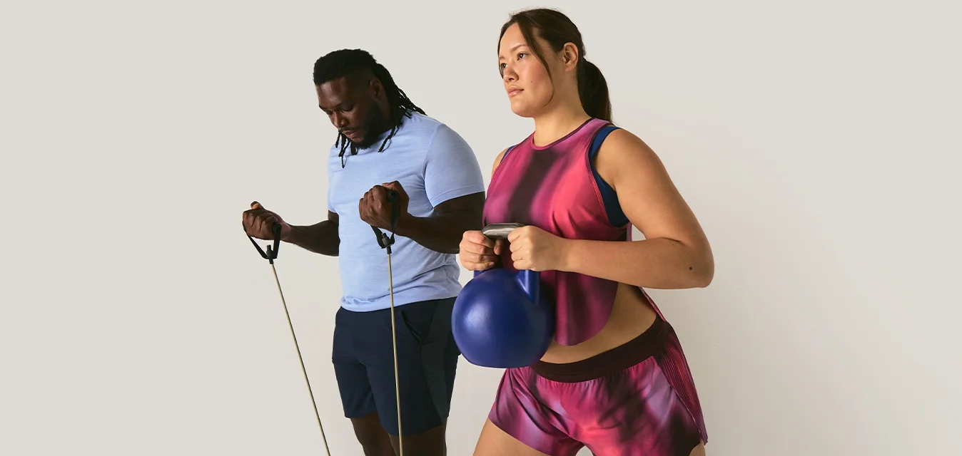 Un uomo si allena con le bande di resistenza mentre una donna tiene un kettlebell blu indossando abbigliamento sportivo rosa su uno sfondo neutro.