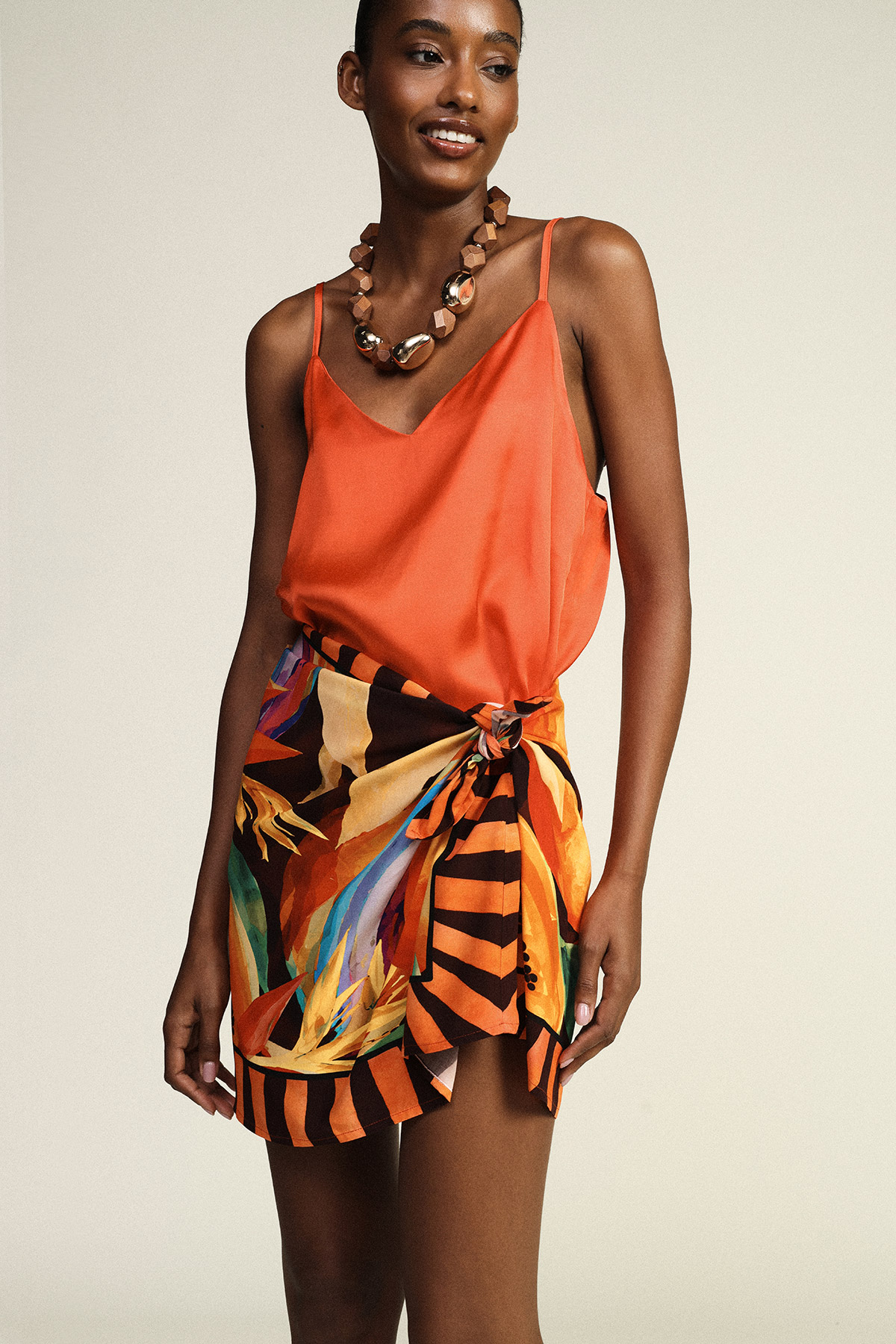 Smiling model wearing orange silk camisole, colourful patterned tie-front wrap skirt and chunky statement necklace