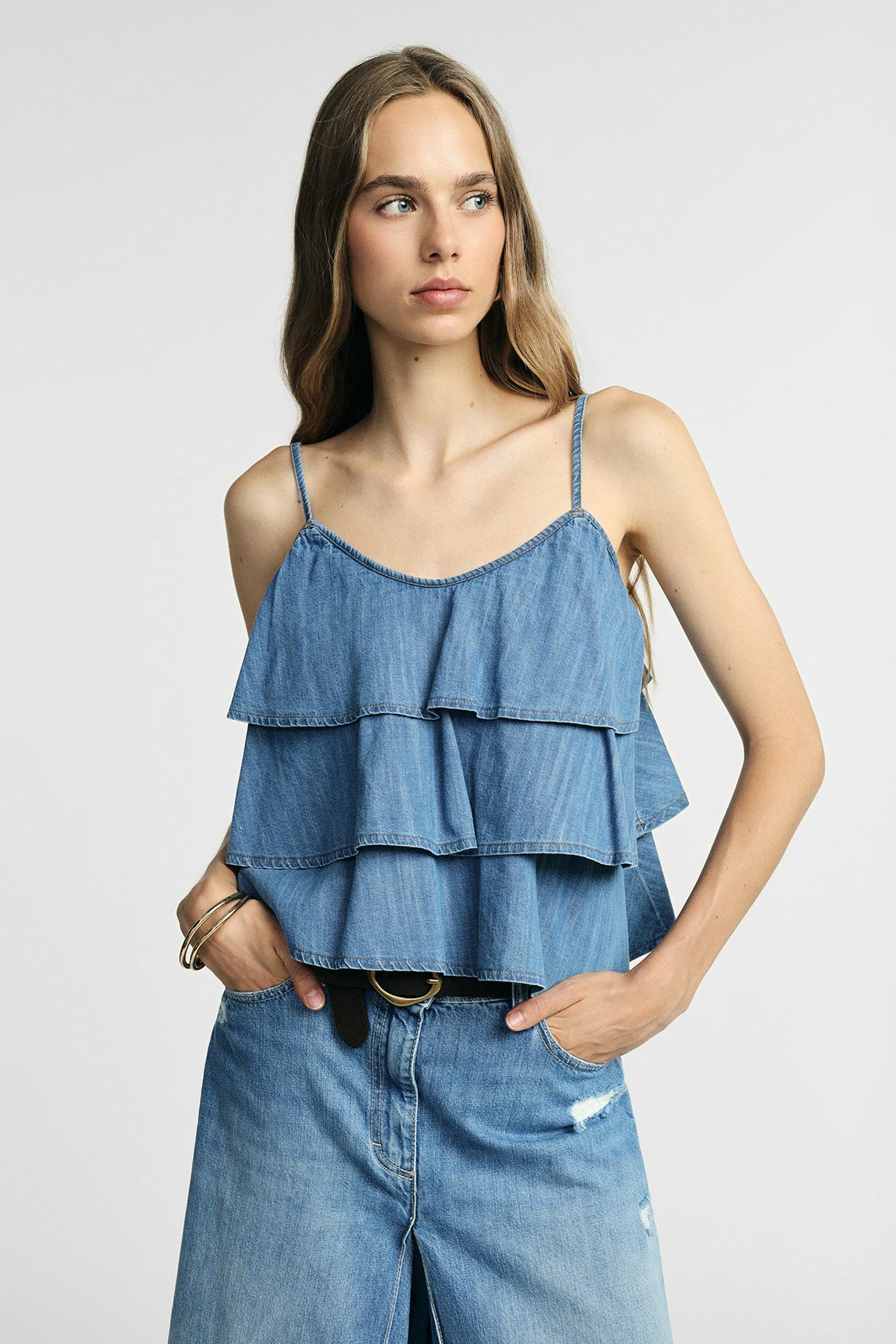 Woman wearing tiered denim cami top and high-waist ripped jeans, hands in pockets against a neutral background