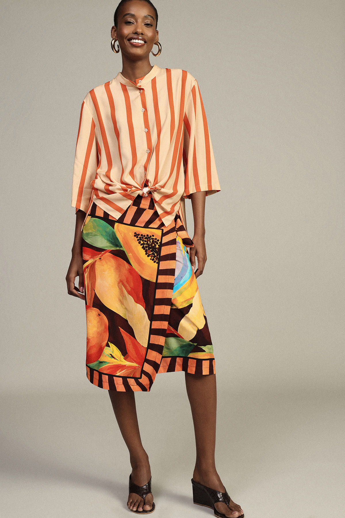 Smiling model in orange striped tie-front blouse and colourful tropical wrap skirt with gold hoops and black wedge sandals