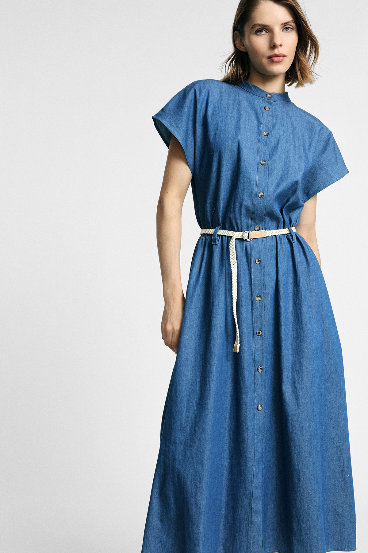 Model wearing blue denim button-front midi dress with short sleeves and rope belt, standing against a plain light background