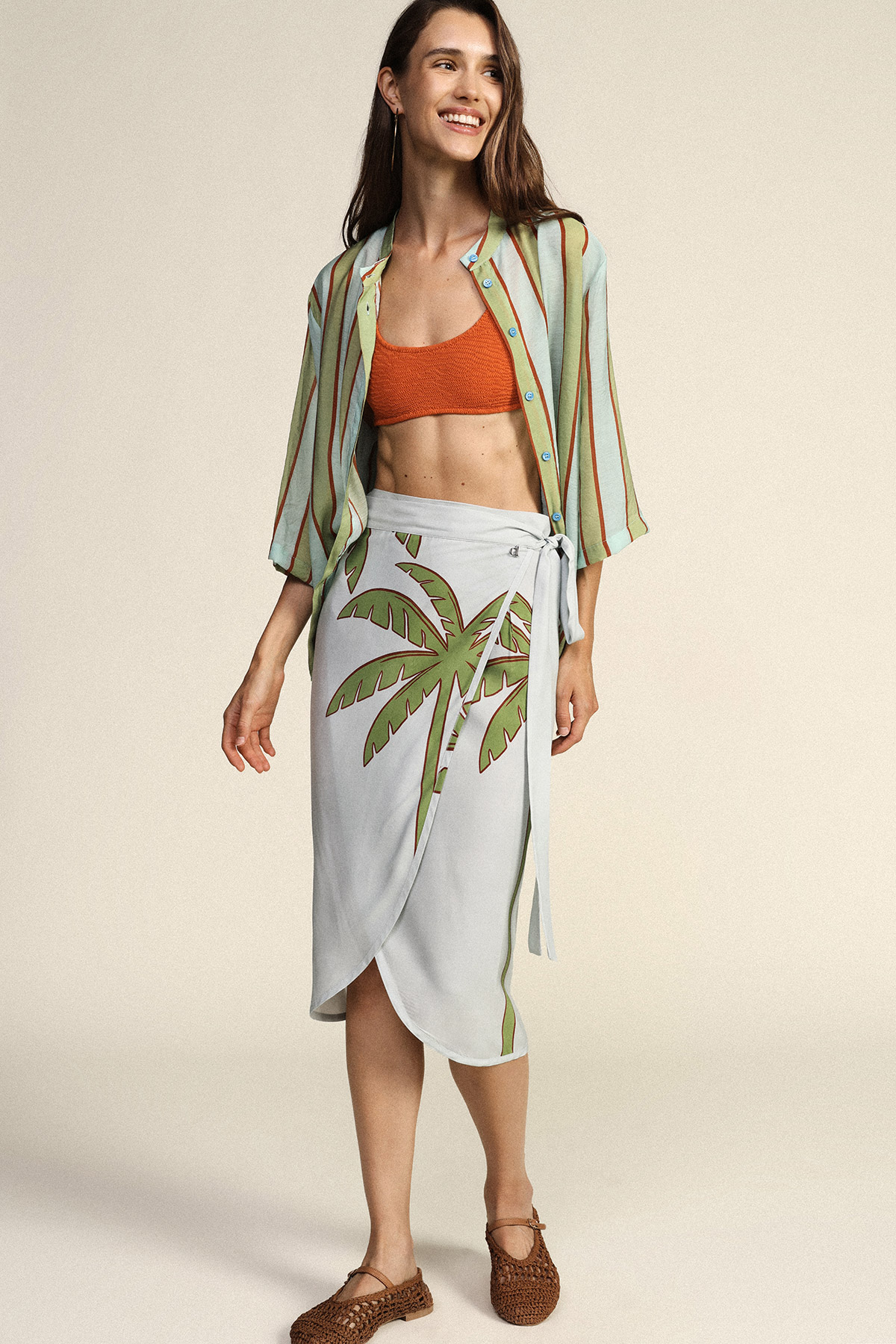 Woman modelling beachwear: orange bikini top, green striped open shirt and white wrap skirt with palm print, woven flats