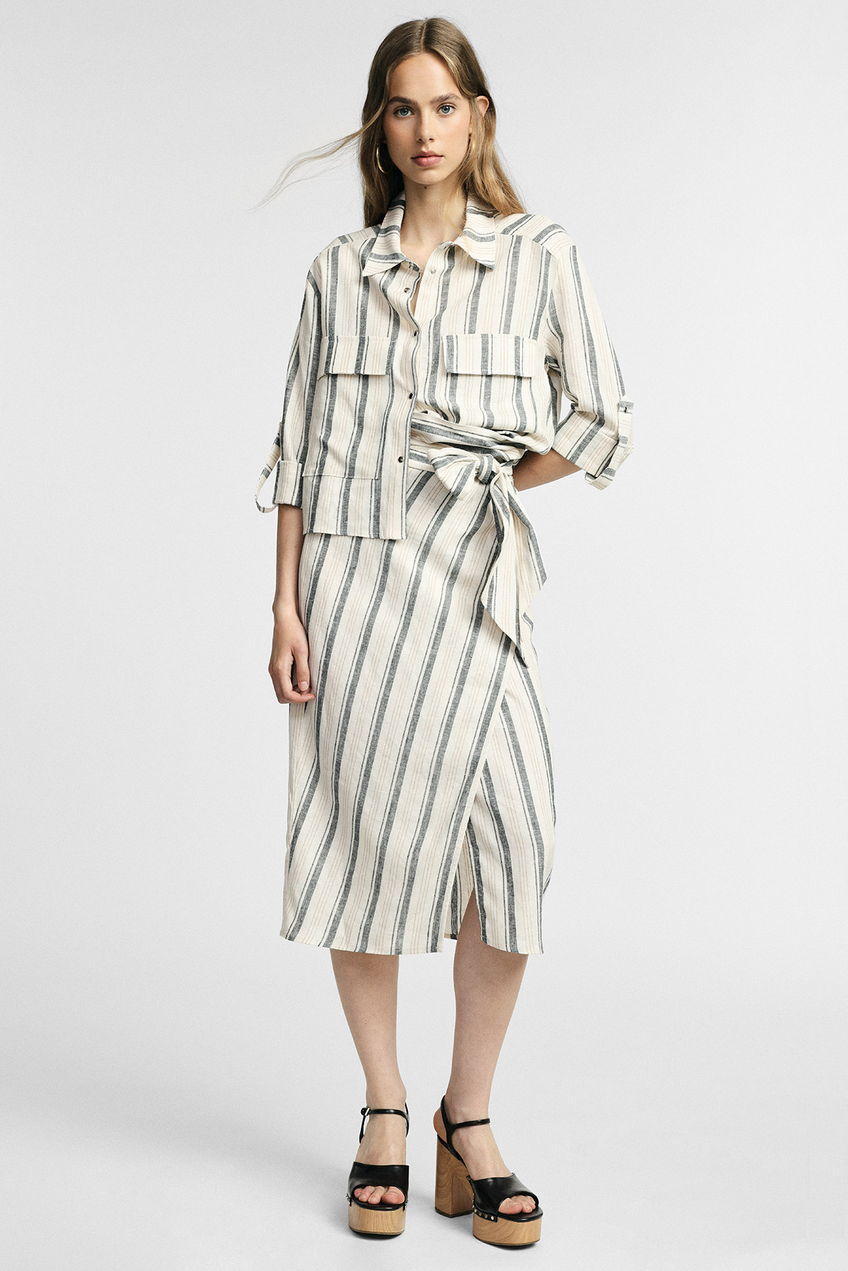 Woman modelling cream and grey vertical striped button-up shirt and matching wrap midi skirt with tie, wearing black platform sandals