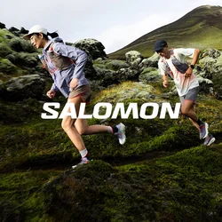 Due corridori trail con abbigliamento sportivo percorrono un pendio roccioso e muschioso con una montagna sullo sfondo.