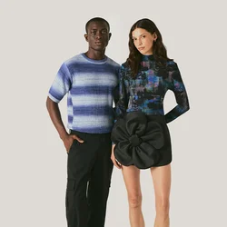 Un uomo con un maglione a righe blu e pantaloni neri sta accanto a una donna che indossa una maglia a maniche lunghe fantasia e una gonna nera con un grande dettaglio floreale.