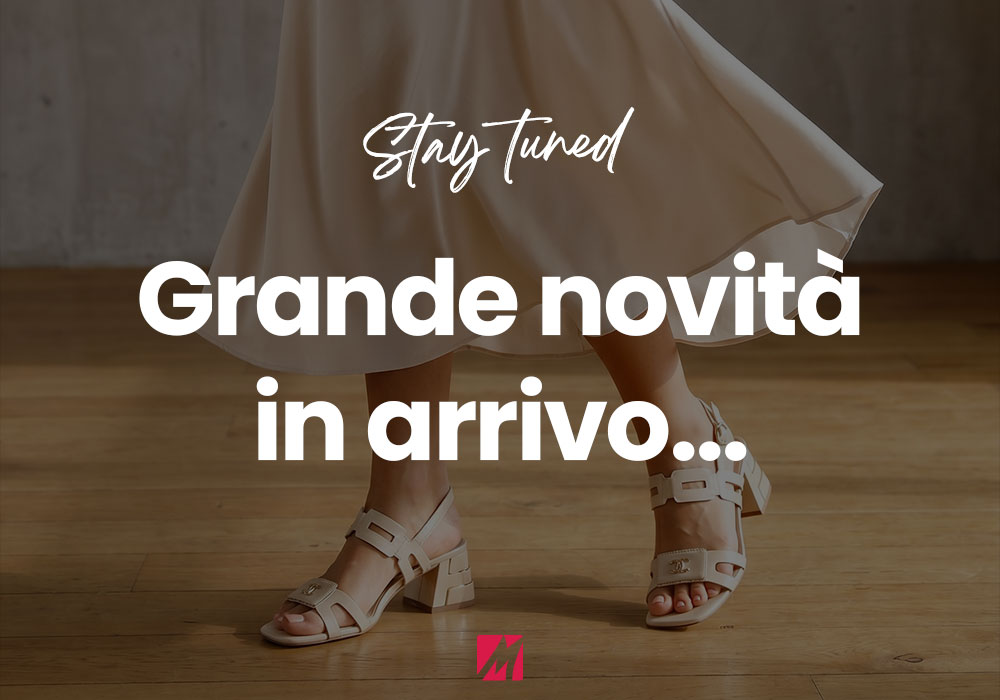 Woman's legs in beige mid-heel sandals and flowing skirt on wooden floor with 'Stay tuned — Grande novità in arrivo...' overlay