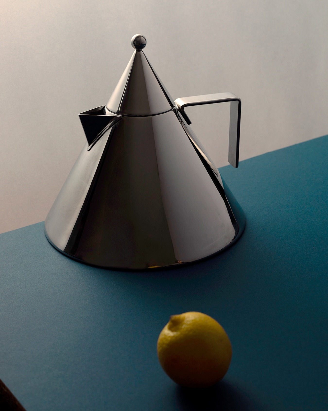 Polished stainless-steel conical teapot with angular handle on teal tabletop, lemon in foreground, minimalist still life