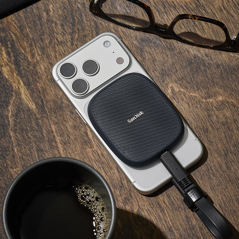 Un SanDisk Phone SSD collegato a uno smartphone poggia su una scrivania di legno accanto a bicchieri e una tazza di caffè.