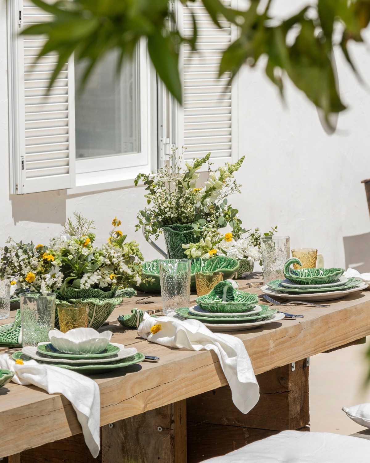 Outdoor wooden dining table set with green ceramic plates, glassware and white napkins, summer wildflower centrepieces by a white shuttered wall