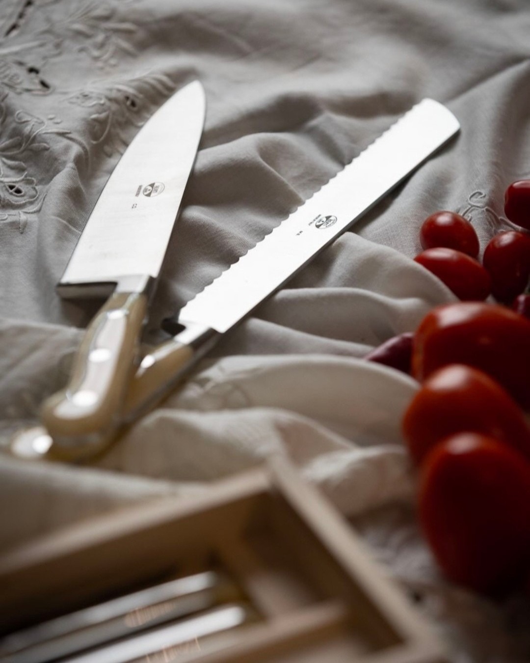 Chef's knife and serrated bread knife on a soft embroidered cloth beside a cluster of ripe cherry tomatoes