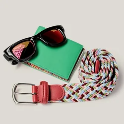 Occhiali da sole neri su un portafoglio verde accanto a una cintura intrecciata multicolore arrotolata con fibbia in pelle rossa su sfondo chiaro.
