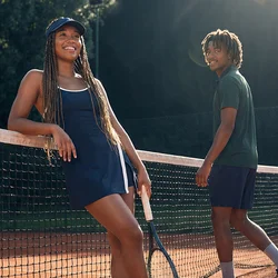 Due giovani stanno su un campo da tennis in terra rossa vicino alla rete, uno appoggiato con una racchetta, luce del sole e alberi sullo sfondo.