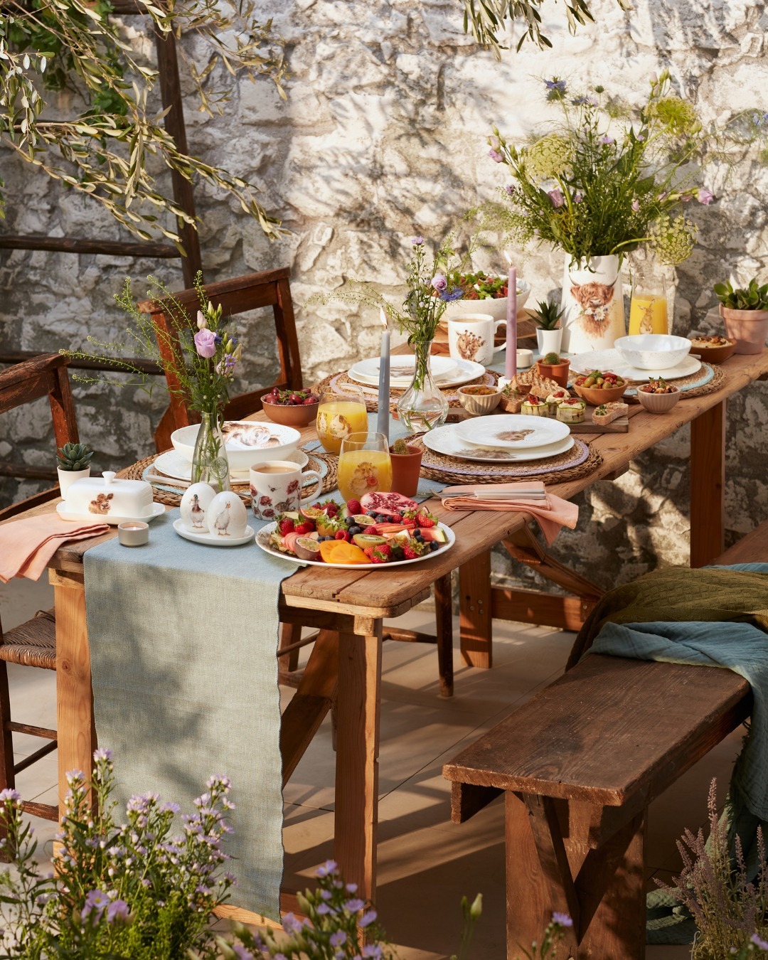 Rustic outdoor wooden table set for brunch with plates, fruit platter, orange juice, candles and wildflower centrepieces
