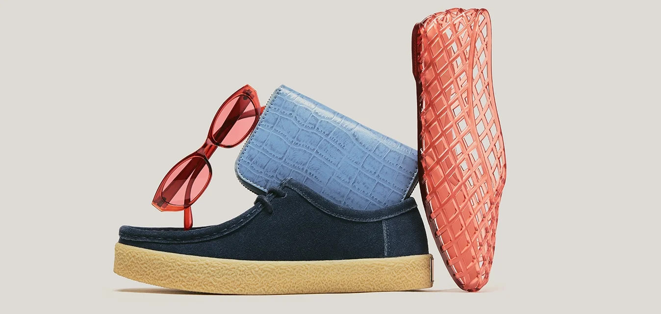 Occhiali da sole rossi, pantofole blu con suola color cuoio e borsa corallo con texture disposte su sfondo beige.