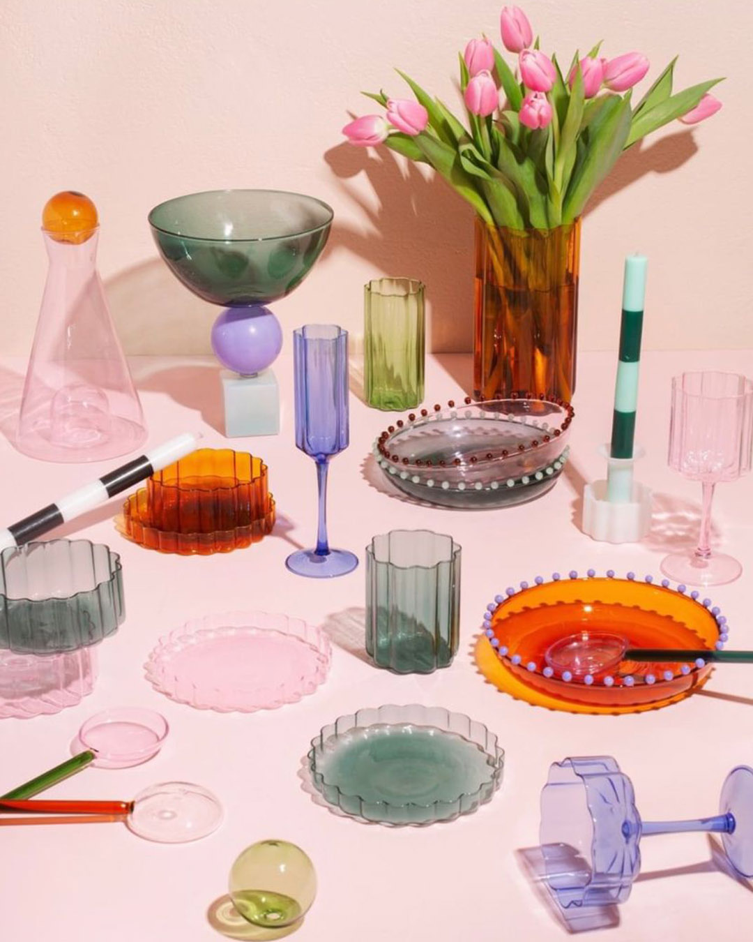 Assorted colourful glassware, plates and bowls arranged on a pale pink table beside a tall amber vase of pink tulips