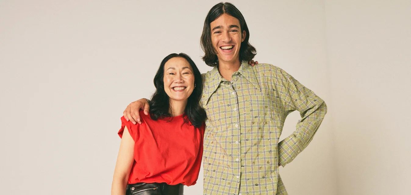 Due persone sorridenti che stanno vicine; una indossa una maglietta rossa e pantaloni neri, l'altra una camicia a quadri verde e jeans chiari.