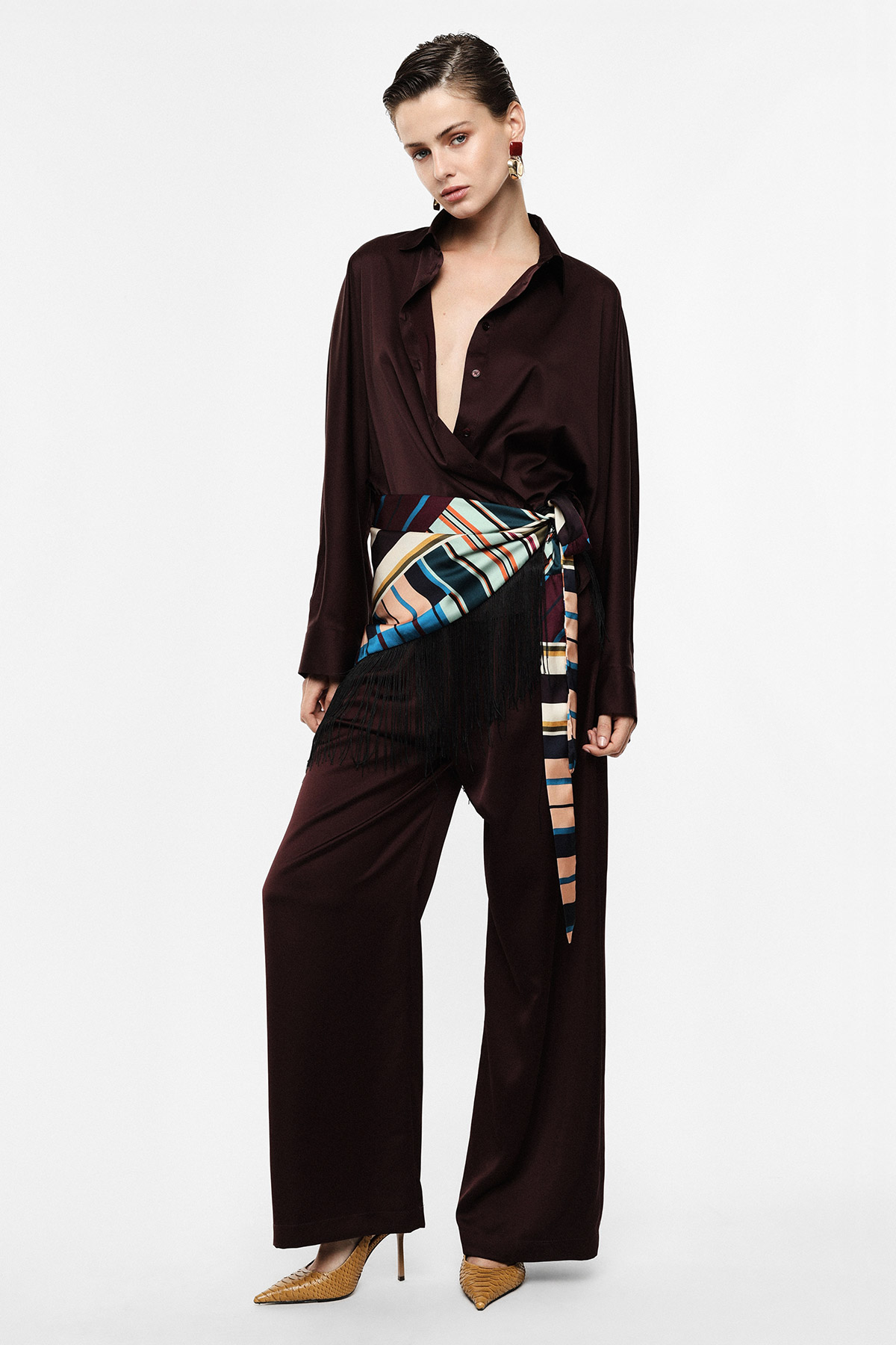 Woman model in burgundy relaxed-fit shirt and wide-leg trousers, striped silk sash with fringe and tan heels — contemporary women's fashion