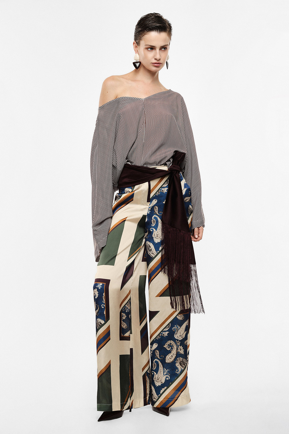 Woman wearing off-shoulder striped blouse and patterned wide-leg trousers with fringed sash belt, studio fashion portrait