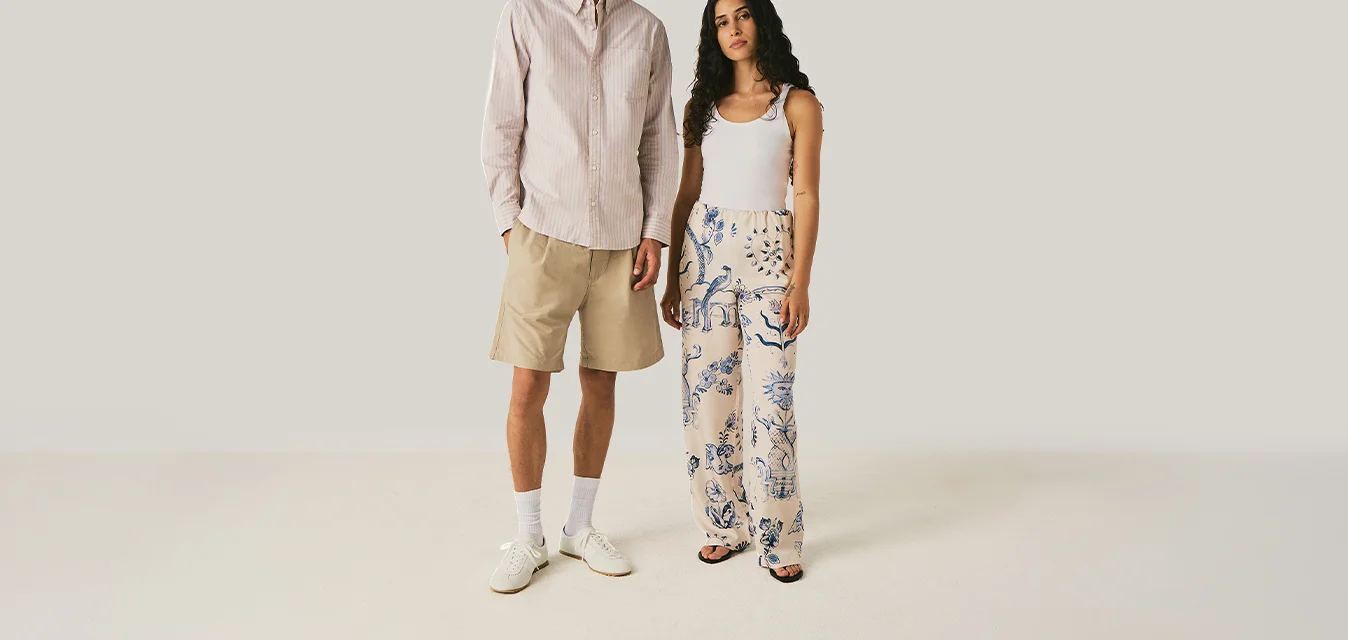 Uomo con pantaloncini beige, camicia bianca, calzini bianchi e scarpe, in piedi accanto a una donna con canotta bianca e pantaloni larghi a fantasia su sfondo uniforme.