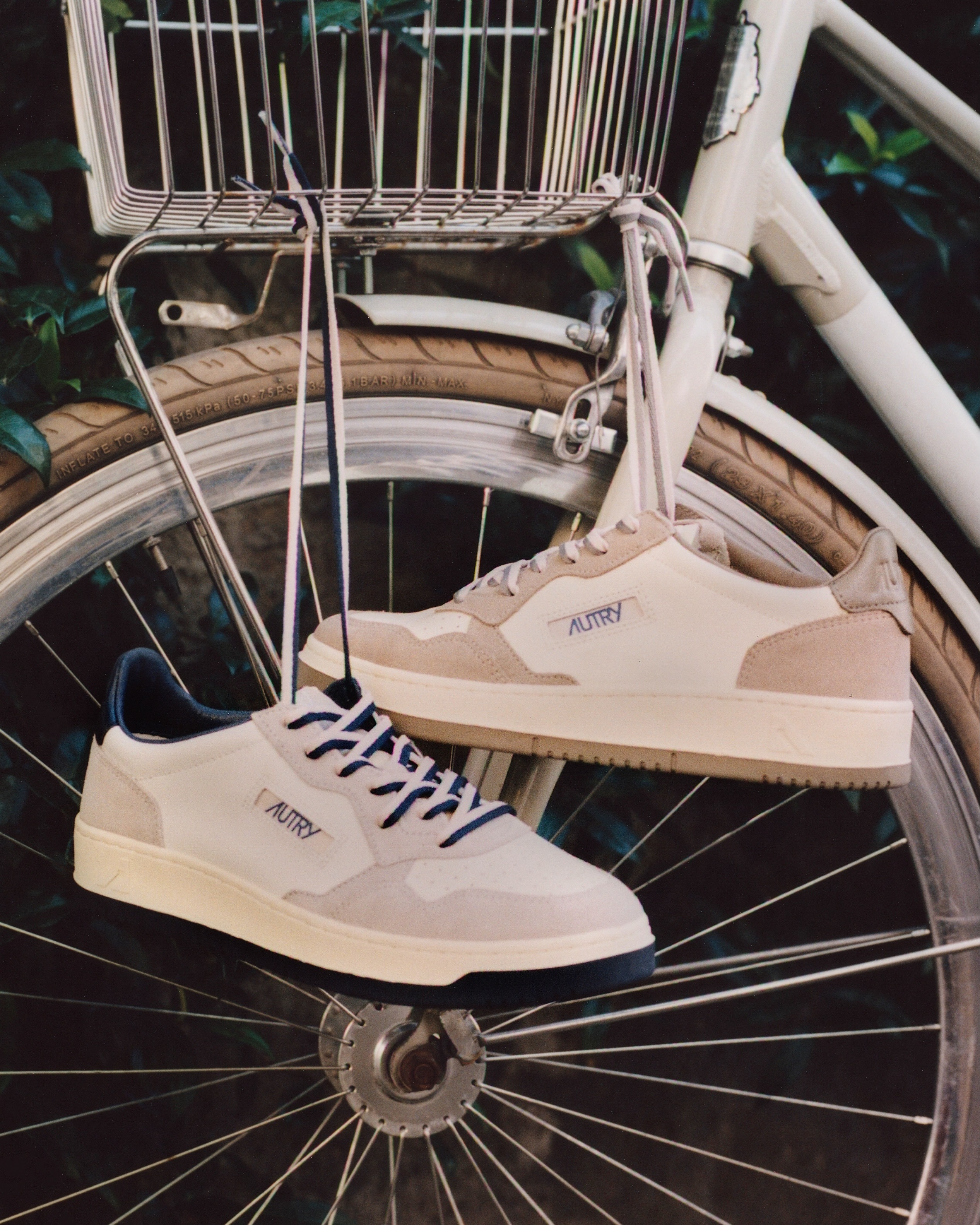 Pair of cream Autry sneakers hanging by their laces from a vintage bicycle wheel and front basket, close-up lifestyle shot