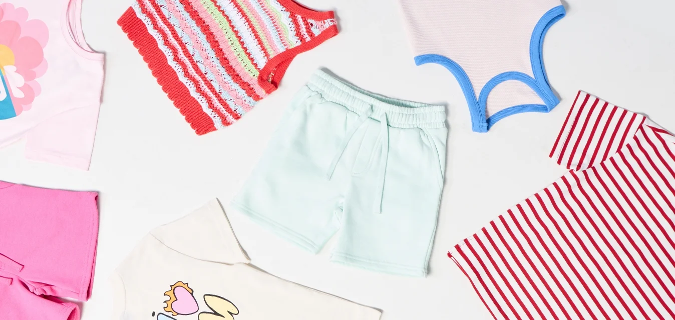 Un assortimento di abbigliamento per bambini include un gilet a righe, pantaloni corti pastello, t-shirt con grafiche e pantaloni corti floreali in vari colori e motivi.