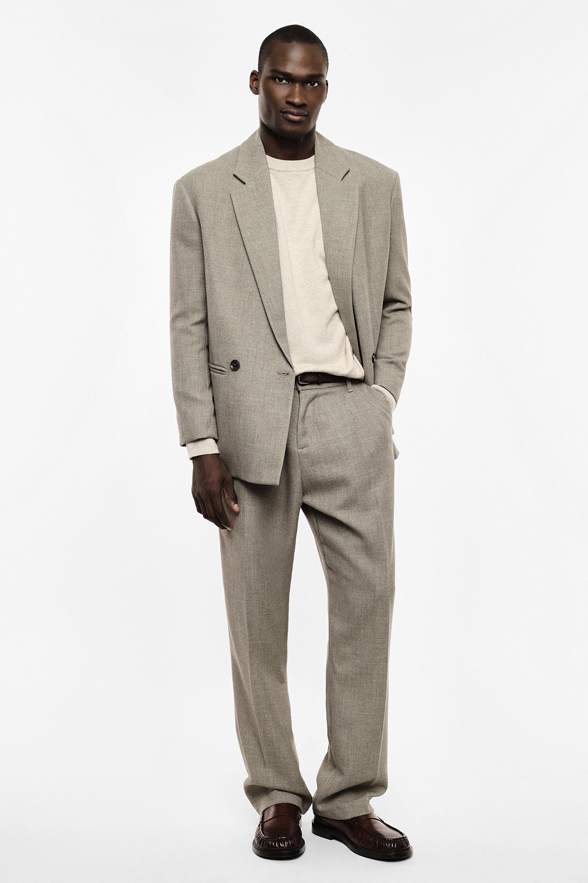 Male model wearing beige relaxed-fit suit with cream sweater and brown leather loafers on white background