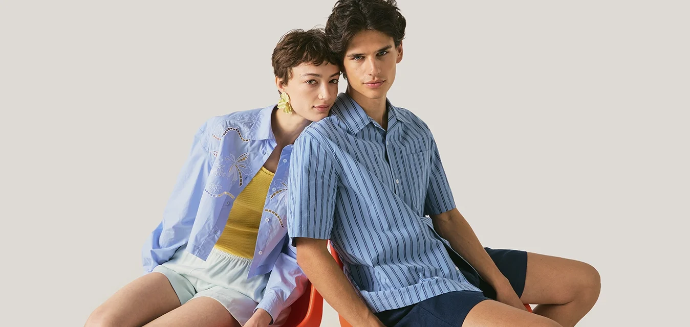 Due persone sedute vicino su una sedia rossa, una indossa una camicia a righe blu e pantaloncini scuri, l’altra una camicia azzurro chiaro con pantaloncini bianchi.